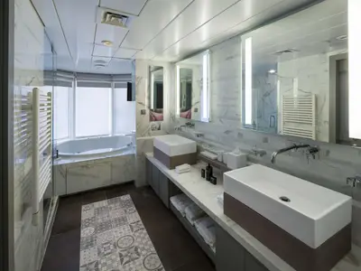 Modernes Bad mit Doppelwaschtisch, beleuchteten Spiegeln und Eckbadewanne vor Fenstern. Wände in Marmoroptik, Boden mit Muster. Handtuchheizkörper links.