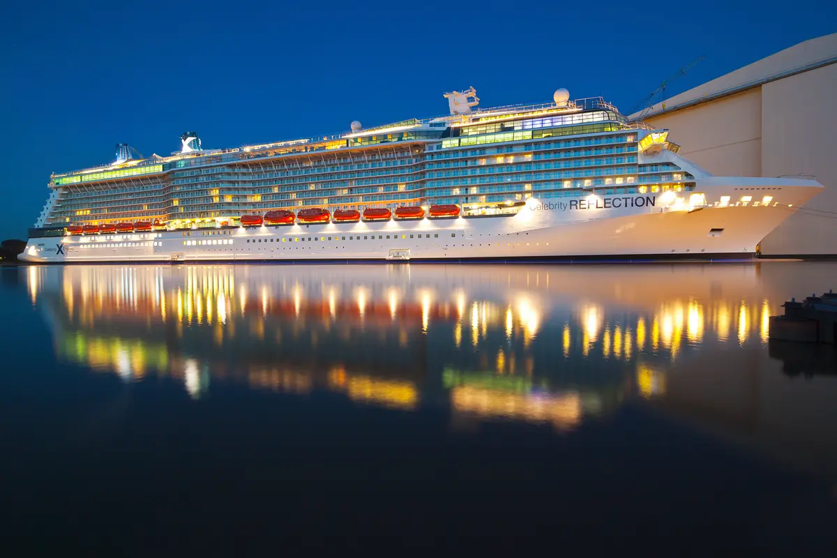 Ein grosses, weisses Kreuzfahrtschiff namens 'Celebrity Reflection' liegt im Hafen bei Nacht. Die Lichter des Schiffes spiegeln sich klar im ruhigen Wasser.