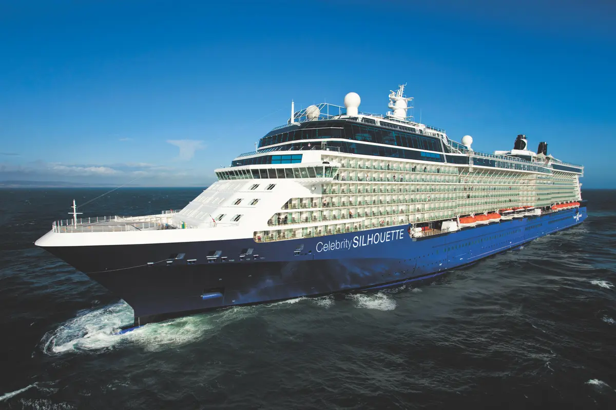 Ein großes, blau-weißes Kreuzfahrtschiff namens 'Celebrity Silhouette' fährt auf dem offenen Meer. Der Bug zeigt nach links, das Schiff erzeugt Bugwellen. Ein strahlend blauer Himmel mit einigen Wolken spannt sich über dem Meer.