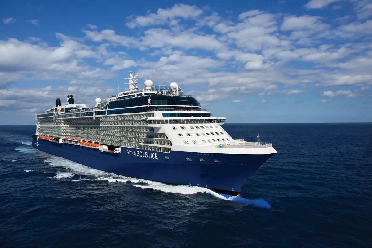 Ein grosses blau-weisses Kreuzfahrtschiff, die Celebrity Solstice, faehrt auf dem offenen Meer unter einem blauen Himmel mit weissen Wolken, Bug voraus.