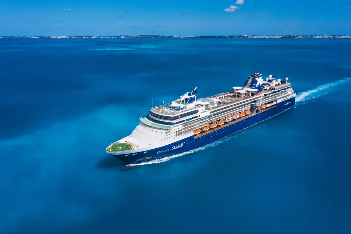 Luftaufnahme des Celebrity Summit Kreuzfahrtschiffes in leuchtend blauem Wasser, die Bugseite mit einem Hubschrauberlandeplatz ist sichtbar. Im Hintergrund ist eine Küstenlinie zu erkennen. Das Schiff hinterlässt eine weisse Kielspur.