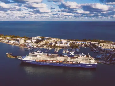 Luftaufnahme des Kreuzfahrtschiffs Celebrity Summit, festgemacht in einem belebten Hafen. Umgeben von Yachten, weißen Gebäuden und Meer unter einem teils bewölkten Himmel.
