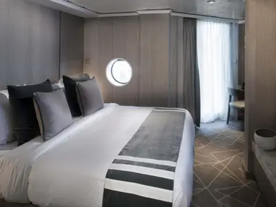Moderne Kabine mit Doppelbett und grau-weißer Bettwäsche. Ein Bullauge, Fenster, Schreibtisch und TV prägen den Raum in Grautönen mit geometrischem Teppich.