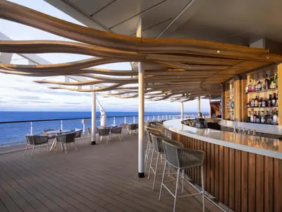 Moderne Außenbar an Deck der Celebrity Summit. Langer Tresen, Barhocker, Sitzbereiche auf Holzdeck. Blaues Meer und heller Himmel im Hintergrund.