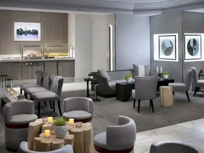 Eine moderne Hotellounge mit einem Buffetbereich auf der linken Seite, mit warmen Speisen und Gebaeck. Auf der rechten Seite sind bequeme Sessel und Sofas in Grautoenen mit Holztischen und gruenen Pflanzen arrangiert. Leuchtende Kunstwerke an der Wand. Der Raum ist hell und einladend.