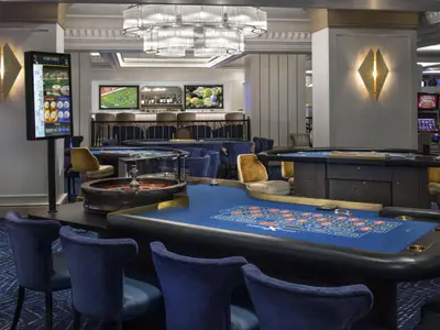 Modernes Casino der Celebrity Summit mit blauen Roulette- und Kartenspieltischen, dazu passende Stühle, eine Bar und digitale Anzeigen.