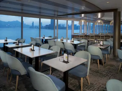 Ein modernes Restaurant an Bord der Celebrity Summit mit zahlreichen Tischen und Stühlen. Durch die Fenster sieht man das Meer und eine Stadt im Abendlicht.