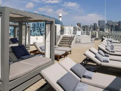 Sonnendeck der Celebrity Summit mit mehreren Reihen von Liegestühlen, einigen Cabanas und einer Treppe. Im Hintergrund ist eine Stadtsilhouette zu sehen.