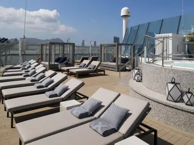 Sonnendeck der Celebrity Summit mit hellen Liegen, Cabanas und einem Whirlpool. Im Hintergrund sind eine Stadtkulisse und ein blauer Himmel zu sehen.