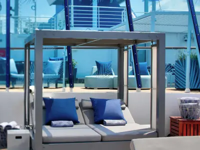Eine moderne Cabana mit zwei grauen Liegen und blauen Kissen auf dem Oberdeck der Celebrity Summit. Im Hintergrund sind weitere Lounge-Möbel und blaue Glaswände sichtbar, die eine entspannte maritime Atmosphäre schaffen.