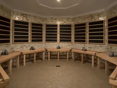 Eine Infrarot-Wärmekabine mit hellbraunen Holzliegen, auf denen graue Handtücher liegen. Die Wände sind mit kleinen Fliesen verkleidet, und an den Wänden befinden sich mehrere dunkle Infrarot-Heizelemente mit Holzrahmen. Der Boden ist mit kleinen braunen Mosaikfliesen ausgelegt.