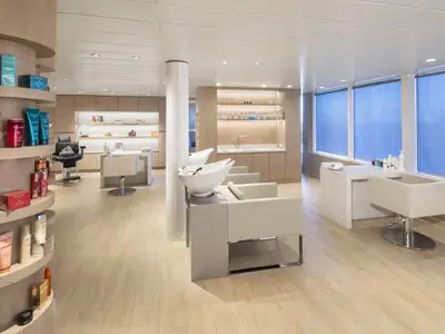 Modern eingerichteter Spa- und Friseursalon auf der Celebrity Summit mit mehreren Styling- und Haarwaschplätzen. Produktregale und große Fenster mit blauen Rollos prägen den hellen Raum.