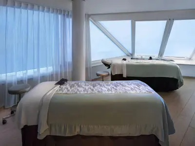 Heller Spa-Behandlungsraum mit zwei Massageliegen und großen Fenstern, die viel Tageslicht hereinlassen.