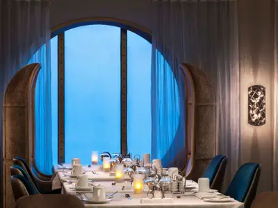 Ein eleganter Restaurantbereich auf der Celebrity Summit mit einem langen, weiß gedeckten Tisch. Durch das große, ovale Fenster ist ein hellblauer Himmel oder das Meer sichtbar. Mehrere Kerzenlichter und blaue Stühle ergänzen die Szene.
