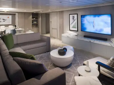 Modernes Wohnzimmer an Bord der Celebrity Summit. Ein graues Ecksofa, Couchtische, großer Flachbildfernseher an einer Wand mit Marmorablage. Im Hintergrund ein Essbereich.