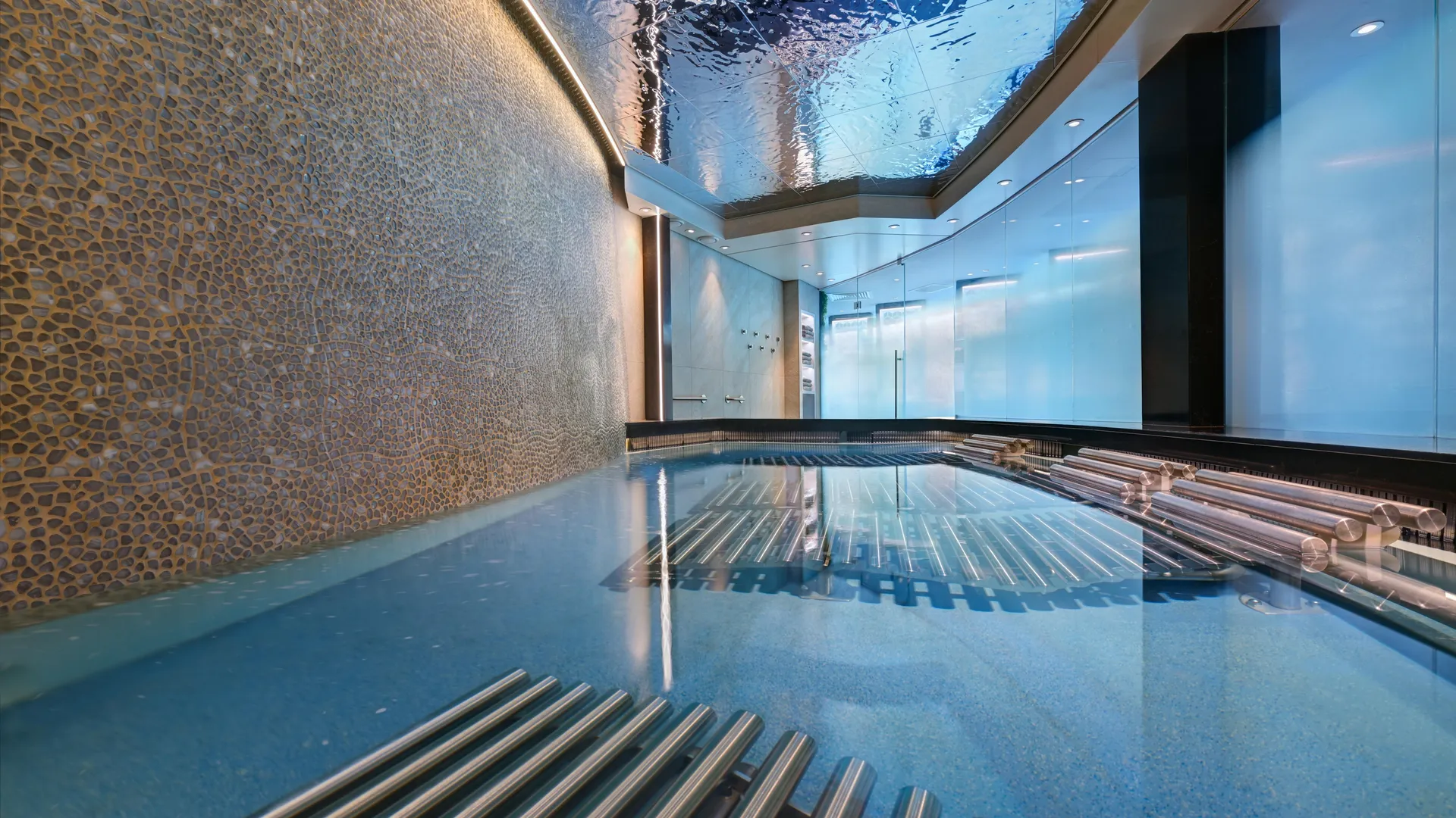 Moderner Innenpool auf der Celebrity Xcel. Blaue Unterwasserliegen im Becken, eine strukturierte Mosaikwand und eine reflektierende Wellendecke. Bläuliches Licht schafft ein beruhigendes Ambiente.