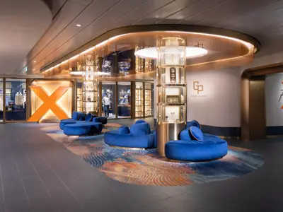 Moderne Lounge der Celebrity Xcel: Blaue Samtsofas auf buntem Teppich. Hohe Vitrinen, großes X-Logo, Ladenfronten mit Kleidung. Warme Beleuchtung, goldene Akzente.