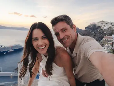 Lachendes Paar in einem Selfie vor einer malerischen Küste mit weißen Gebäuden und dem Kreuzfahrtschiff Celebrity Xcel im blauen Meer.