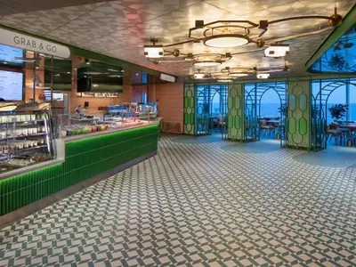 Blick in den Grab & Go Bereich auf der Celebrity Xcel mit grüner Theke, Deckenleuchten und großen Fenstern mit Meerblick. Der Boden hat ein geometrisches Fliesenmuster.