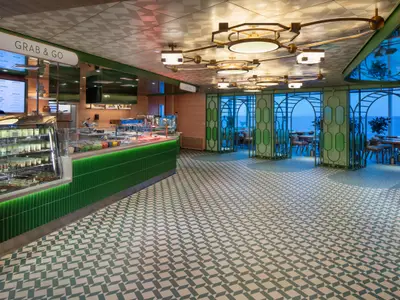 Innenansicht des Grab & Go Restaurants auf der Celebrity Xcel. Grüne Theke, Meerblick durch Bogenfenster, gemusterter Boden, goldene Deckenleuchten.