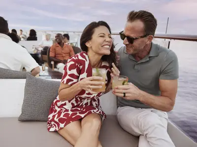 Ein lachendes Paar sitzt mit Cocktails auf dem Außendeck der Celebrity Xcel. Im Hintergrund sind weitere Gäste und das Meer zu sehen, was entspannte Stimmung vermittelt.