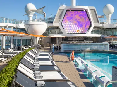 Das sonnige Pooldeck der Celebrity Xcel zeigt Liegestühle, orangefarbene Sonnenschirme, einen blauen Pool, eine Poolbar und eine große LED-Wand.
