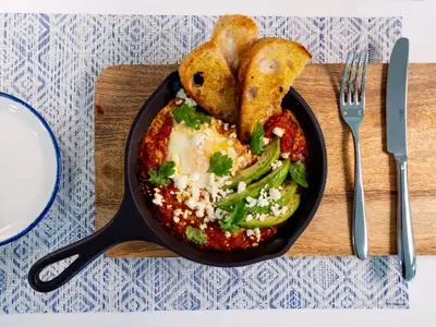 Ein appetitlich angerichtetes Gericht in einer schwarzen gusseisernen Pfanne, bestehend aus Spiegelei, roter Tomatensauce, geschnittener Avocado und zerbröseltem Feta, garniert mit Koriander. Dazu zwei Scheiben geröstetes Brot.
