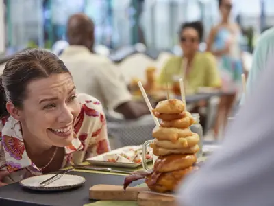 Eine Frau in einem gemusterten Hemd lacht und schaut begeistert auf einen hoch gestapelten Burger mit vielen Zwiebelringen. Im Hintergrund sind undeutlich weitere Gäste zu sehen.