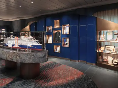 Ein detailliertes Modell der Celebrity Xcel in einer ovalen Glasvitrine steht im Zentrum eines blauen Ausstellungsraums. An der Wand sind weitere Vitrinen mit nautischen Gegenständen zu sehen.