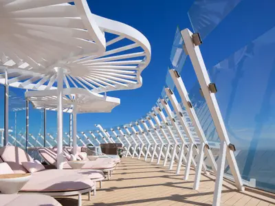 Sonnendeck der Celebrity Xcel mit weißen, fächerförmigen Sonnenschirmen und rosa Liegen auf Holzdeck. Eine geschwungene Glaswand mit weißen Streben ermöglicht weiten Blick unter blauem Himmel.