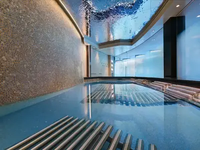 Moderner Innenpool auf der Celebrity Xcel. Blaue Unterwasserliegen im Becken, eine strukturierte Mosaikwand und eine reflektierende Wellendecke. Bläuliches Licht schafft ein beruhigendes Ambiente.
