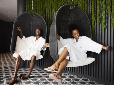Ein lächelndes Paar in weißen Bademänteln sitzt entspannt in zwei dunklen Hängesesseln in einer modernen Spa-Lounge. Im Hintergrund sind begrünte Wände zu sehen.