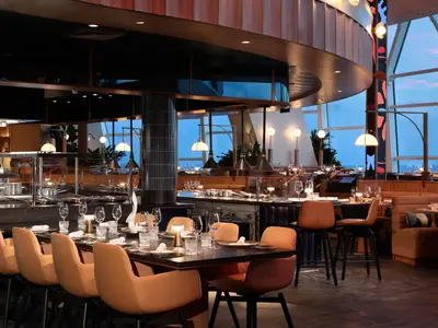 Blick in ein modernes Restaurant an Bord der Celebrity Xcel mit langer Tafel, offener Küche und bodentiefen Fenstern, die den Blick auf das Meer freigeben.