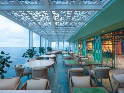 Restaurant auf der Celebrity Xcel mit Meerblick. Hohe Fenster, helle Möbel, grüne Akzente und eine dekorative Decke charakterisieren den modernen Innenraum.