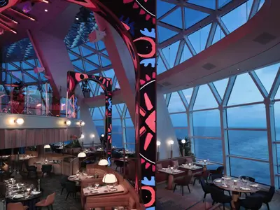 Restaurant der Celebrity Xcel mit Meerblick. Esstische auf mehreren Ebenen. Leuchtende Säulen, eine Bühne mit Schlagzeug.
