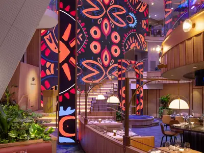 Innenansicht eines mehrstöckigen Restaurants namens "Mosaic" mit Tischen und Stühlen. Eine große Wand ist mit einem farbenfrohen, abstrakten Muster in Orange, Rot und Blau auf schwarzem Grund verziert. Elegante Leuchten und Holzverkleidungen ergänzen das moderne Design.