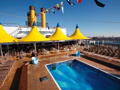Der einladende Poolbereich an Bord der Costa Deliziosa mit gelben Sonnenschirmen, Liegestühlen und Blick auf die Skyline im Hintergrund.