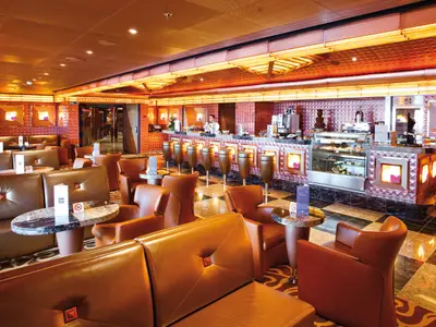 Blick in die elegante Bar Sugar der Costa Deliziosa. Ein Barkeeper bereitet Getränke zu. Zahlreiche braune Ledersofas und Sessel laden zum Verweilen ein. Die Theke bietet Süßspeisen und einen Schokoladenbrunnen.