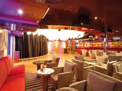 Blick in die elegante Piano Bar Excite an Bord der Costa Deliziosa. Zu sehen sind eine Bühne, ein großer Kronleuchter und viele bequeme Sessel mit Tischen.