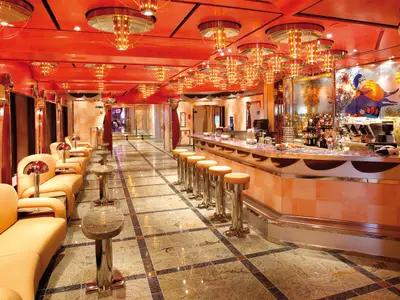 An elegant bar on the Costa Deliziosa with a long counter and bar stools, plush beige sofas, and striking red chandeliers. The Vanilla Bar features artistic glass panels. No people visible.