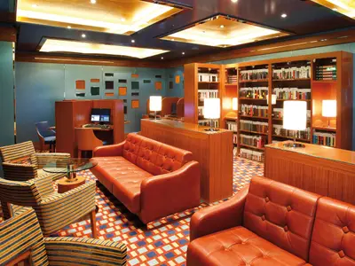 Blick in die Bibliothek der Costa Deliziosa mit orangen Ledersofas, gestreiften Sesseln, Holzregalen voller Buecher und Computern. Ein gemuetlicher Ort zum Entspannen.
