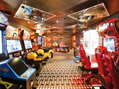 Ein lebhaftes Spielzimmer der Costa Deliziosa mit vielen Arcade-Automaten, Motorrad-Simulatoren und einem Airhockey-Tisch. Der Raum ist hell beleuchtet und hat einen karierten Boden.