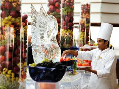 Ein Koch in weißer Uniform serviert frisches Obst am reichhaltig verzierten Buffet an Bord der Costa Deliziosa.