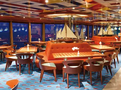 Ein stilvolles Restaurant an Bord der Costa Deliziosa mit dunklen Holztischen und orangefarbenen Sitzgelegenheiten. Im Hintergrund sind durch große Fenster Schiffsmodelle zu sehen.
