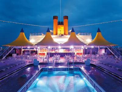 Abendstimmung auf dem Acqua Regina Lido der Costa Deliziosa. Beleuchteter Pool, Liegestühle, Bar unter gelben Pavillons und orange Schornsteine.