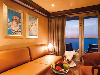 Balkonsuite der Costa Deliziosa: Ledersofa, Kunstwerke, Tisch mit Sekt und Obst. Durch die Tür ist der Balkon mit Stühlen und das Meer bei Abenddämmerung zu sehen.