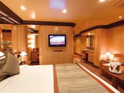 Eine geräumige Suite an Bord der Costa Deliziosa, hell beleuchtet. Im Vordergrund ein grosses Doppelbett mit gemusterten Kissen. Links ein Spiegel und ein Schminktisch, rechts ein Sitzbereich mit Sofa, Couchtisch und Champagner. Ein Flachbildfernseher ist an der Wand montiert.