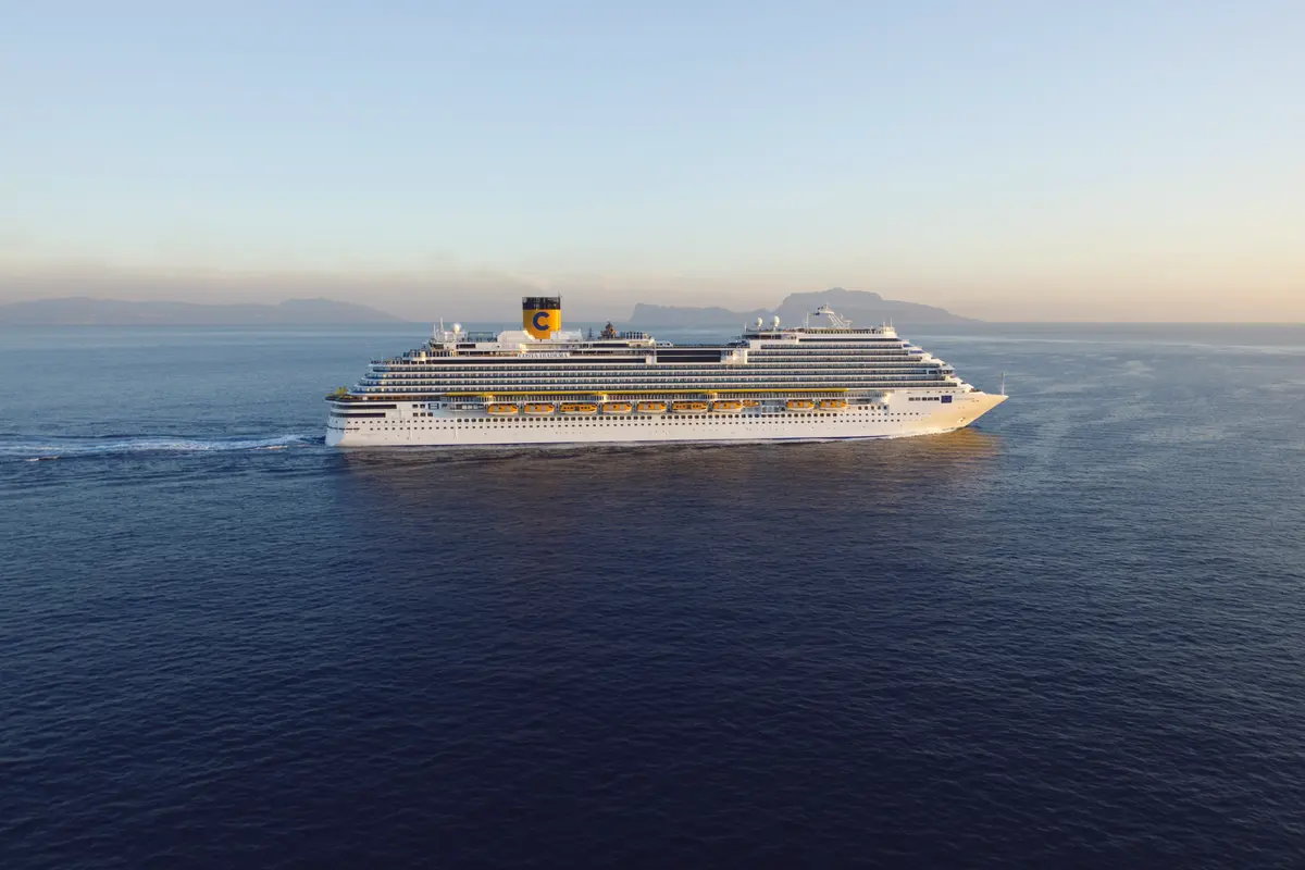 Die weisse Costa Diadema mit gelbem Schornstein fährt auf dem blauen Meer. Sie zieht eine weisse Heckwelle. Berge im Hintergrund unter hellem Himmel.