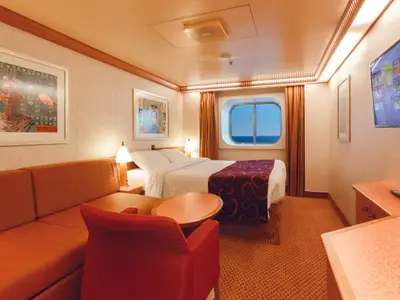 Helle Aussenkabine an Bord der Costa Diadema mit grossem Bullaugenfenster zum Meer. Sie zeigt ein Doppelbett, ein oranges Sofa, einen Tisch und einen Fernseher.
