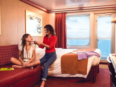 In einer hellen Balkonkabine der Costa Diadema flechtet eine Mutter ihrer lachenden Tochter die Haare. Die Tochter sitzt auf dem roten Sofa, die Mutter auf dem Bett. Das Meer ist durch das Fenster sichtbar.
